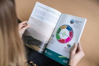 The image shows a person reading a report focused on the Sustainable Development Goals (SDGs). The open magazine features a colorful representation of the SDGs, highlighting topics such as environment, social aspects, and corporate governance.