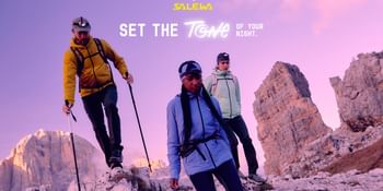 Three hikers with headlamps and trekking poles explore rocky alpine terrain at dusk. They are wearing colorful outdoor gear by Salewa. In the background, the mountains and sky glow in warm purple and pink hues.