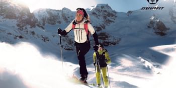 Two ski mountaineers ascend through deep snow under bright sunlight, surrounded by snowy alpine peaks. The person in front wears Dynafit gear.