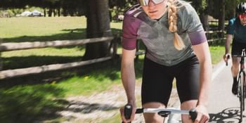 Eine fokussierte Radfahrerin in sportlicher Kleidung fährt auf einer sonnigen Landstraße, gefolgt von einem weiteren Radfahrer.