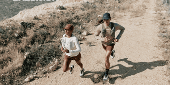 Two female runners are running on a dry trail overlooking a city. The white Salomon logo is visible in the upper left corner.