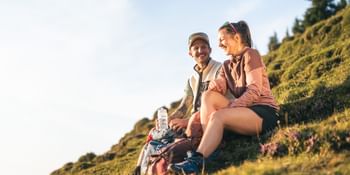Zwei Personen sitzen auf einem grasbewachsenen Hang und lachen miteinander. Neben ihnen liegen Rucksäcke und eine Trinkflasche, im Hintergrund sind Hügel und Bäume im warmen Sonnenlicht zu sehen.