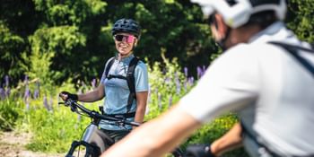 Eine Person mit Helm, Sonnenbrille und Rucksack sitzt auf einem Mountainbike und lächelt zu einer anderen Person, die im Vordergrund unscharf zu sehen ist. Im Hintergrund stehen dichte grüne Bäume und blühende Pflanzen entlang eines Waldweges.