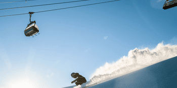 Skifahrer beim hinunterfahren auf der Piste mit einem Sessellift über ihm und schönem Wetter.