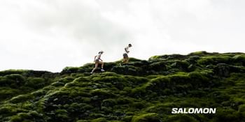 Das Bild zeigt zwei Trailrunner, die über eine mit leuchtend grünem Moos bewachsene Felslandschaft laufen, während der Himmel über ihnen hell und wolkig erscheint; das „Salomon“-Logo ist dezent in der unteren rechten Ecke platziert.