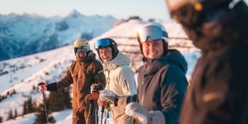 Vier Skifahrer stehen mit Helm, Skibrille und Skistöcken zusammen auf einer verschneiten Piste und lächeln. Im Hintergrund sind schneebedeckte Berge im warmen Morgenlicht zu sehen.