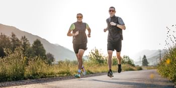 Zwei Läufer joggen nebeneinander auf einem asphaltierten Weg durch eine alpine Landschaft bei tief stehender Sonne.