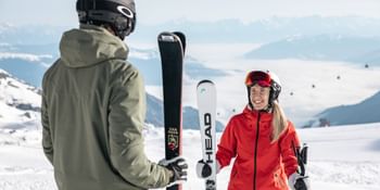 Eine Frau in roter Skiausrüstung und ein Mann in grüner Skiausrüstung unterhalten sich auf der Piste, beide halten ihre Skier.