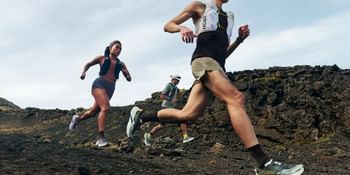 Three trail runners running across a rocky mountain trail wearing Hoka trail running shoes and hydration vests.