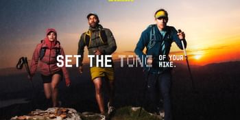 Three people wearing backpacks and outdoor clothing walk along a grassy, rocky mountain path as the sun sets on the horizon. The image includes the text “SET THE TONE OF YOUR HIKE.” and the logo “SALEWA”.