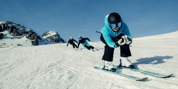 A skier wearing a turquoise jacket, black helmet and goggles descends a snowy slope in a tuck on HEAD skis. Two other skiers follow, with rocky mountain peaks and a blue sky in the background.