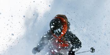 Peak Performance AW 25 Ein Skifahrer in einer dunklen Jacke mit oranger Kapuze fährt durch tiefen Pulverschnee. Schnee wirbelt intensiv um ihn herum und verdeckt teilweise die Sicht.