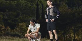 Two trail runners pause on a gravel road in a scenic landscape, wearing Salomon running apparel and trail shoes with forested hills in the background.