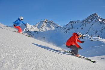 two skiers are ridinge on a downhill tracks, mountains in the background