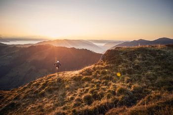 ein Trailläufer befindet sich entlang eines Feldweges auf einem Berghang, im Hintergrund der Sonnenuntergang