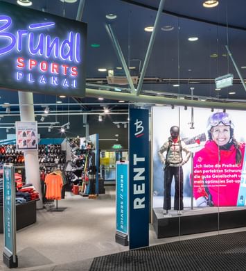 Das Bild zeigt den Eingang eines Bründl Sports Shops in der Planai mit einem großen, leuchtenden Bründl Sports Schild. Im Inneren sind verschiedene Wintersportprodukte wie Kleidung und Ausrüstung ausgestellt, und es gibt einen Bereich für Verleih und Service. Ein großes Werbeposter zeigt eine lächelnde Skifahrerin.