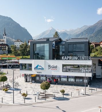 Das Kaprun Center des Kitzsteinhorns vor einer alpinen Bergkulisse. Im Gebäude befindet sich ein Bründl Sports Shop; sichtbar sind unter anderem die Schriftzüge „KAPRUN CENTER“, „Bründl Sports“, „Kitzsteinhorn“, „Tickets“ und „Ski Depot“.