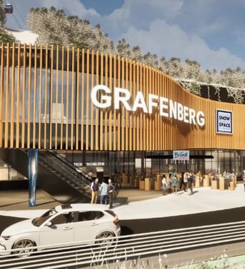 A modern building with a curved wooden facade and the text “GRAFENBERG” stands in front of snow-covered trees. Several people stand near the entrance, a car is passing by, and signs reading “Flachau Snow Space Salzburg,” “Bründl Sports,” and “Ticketschalter” are visible, while a cable car runs above.