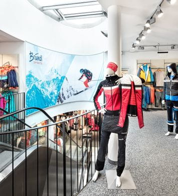 Bründl Sports Saalbach Life.Style - Innenansicht - Rechts im Bild Skitourenoutfits, in der Mitte eine Rolltreppe ins untere Stockwerk und Ski Textil für Kinder links