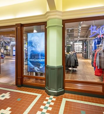 Zu sehen ist die Glasfront des Shops Bründl Sports McArthurGlen Designer Outlet Salzburg mit sichtbarem „Bründl Sports“ Logo im Schaufenster. Im Inneren sind Sportbekleidung, Ski, Helme und Zubehör zu sehen; auf einem Bildschirm steht der Text „#BRÜNDLFAN“, im Hintergrund ist außerdem „kassa“ erkennbar.