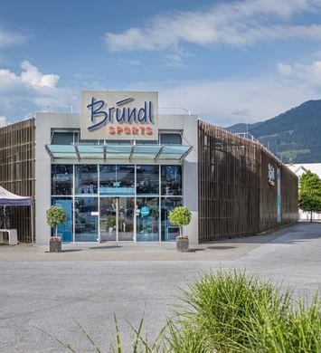 Außenansicht der Bründl Sports Filiale in Saalfelden. Vor dem modernen Geschäftsgebäude mit großem Bründl Sports Logo stehen mehrere Fahrräder unter zwei weißen Zelten. Im Hintergrund sind Berge, Bäume und ein teilweise bewölkter Himmel zu sehen. Sichtbarer Text: „Bründl Sports“ und „Einfahrt“.