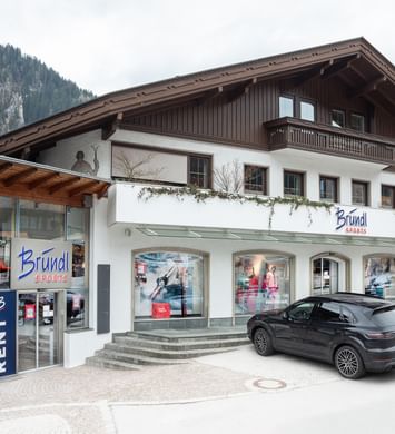 Das Bild zeigt einen Bründl Sports Shop in einem alpenländischen Gebäude mit Schaufenstern, einem Auto davor und Bergen im Hintergrund.