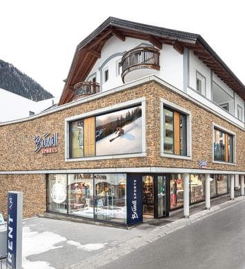 Bründl Sports Ischgl Zentrum mit Steinfassade, großen Schaufenstern und Bründl Sports Logos bei Tageslicht. Vor dem Shop steht ein „RENT“-Schild; Schneereste säumen die Straße und schneebedeckte Berge liegen im Hintergrund.