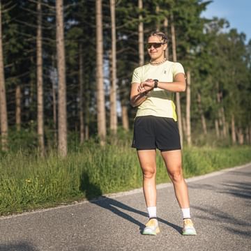 Female runner stands on a paved path at the edge of a forest, looking at her sports watch in sunny conditions.