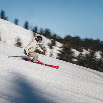 A skier in light-colored ski clothing descends a groomed slope on red skis. The blurred background emphasizes the skier’s speed and movement.