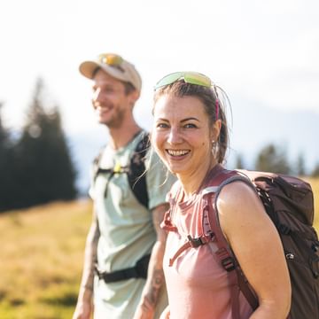 Eine Frau mit Rucksack und Sonnenbrille auf dem Kopf lächelt in die Kamera, während ein Mann mit Kappe und Rucksack leicht unscharf im Hintergrund steht. Sie befinden sich auf einer sonnigen Wiese mit Bäumen und Bergen im Hintergrund.