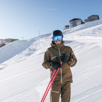 Ein lächelnder Skifahrer in brauner Skibekleidung, schwarzem Helm und blau verspiegelter Skibrille hält rote Skistöcke. Im Hintergrund sind eine präparierte Skipiste, verschneite Hänge und Berggebäude unter blauem Himmel zu sehen.