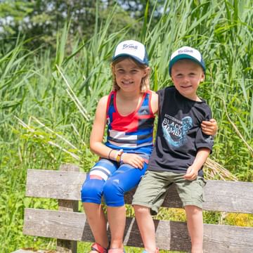 Zwei Kinder mit Bründl Sports-Kappen sitzen lächelnd auf einer Bank inmitten hoher Gräser und genießen die Natur.