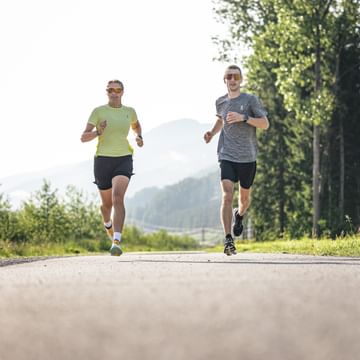Two runners jog on a paved path surrounded by forest and mountains in sunny conditions.