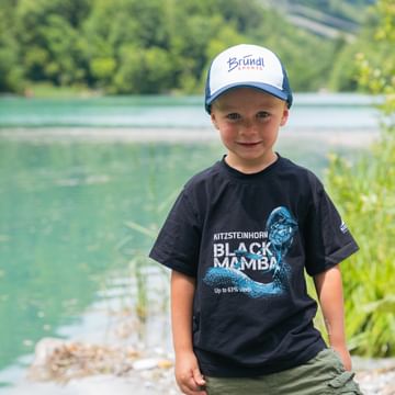 Ein kleiner Junge steht lächelnd am Ufer eines türkisfarbenen Sees, trägt eine Bründl Sports-Kappe, umgeben von grüner Natur.