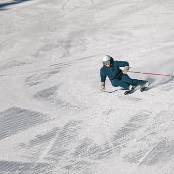 A skier in blue ski clothing leans low while skiing across a groomed slope, spraying snow into the air. Numerous ski and grooming tracks are visible on the snow.