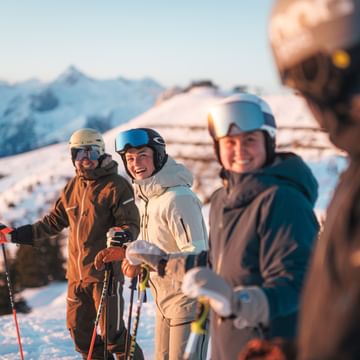 Vier Skifahrer stehen mit Helm, Skibrille und Skistöcken zusammen auf einer verschneiten Piste und lächeln. Im Hintergrund sind schneebedeckte Berge im warmen Morgenlicht zu sehen.