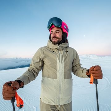 A smiling skier wearing a pink helmet and goggles stands on a snowy slope at sunrise, with mountain peaks in the background.