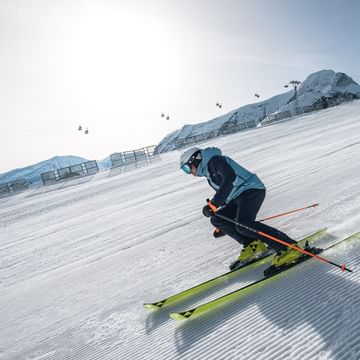 Ein Skifahrer in blauer Skiausrüstung fährt eine frisch präparierte Piste hinunter. Im Hintergrund sind schneebedeckte Berge, ein Seilbahnträger und Gondeln zu sehen.