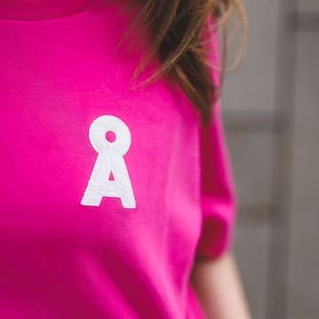 Close-up of a pink shirt with a white letter “A” on the chest. Part of the hair and a necklace are visible, with a blurred stone wall in the background.