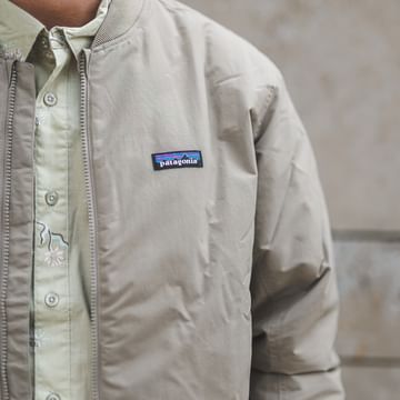 Close-up of a person’s upper body wearing a beige jacket with a small “patagonia” logo visible. Underneath, a light-colored shirt with a subtle pattern can be seen, with a stone wall in the background.