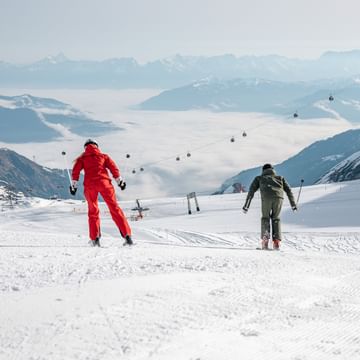 Ein Mann in grüner und eine Frau in roter Skiausrüstung fahren gemeinsam eine verschneite Piste hinunter. Im Hintergrund sind eine Berglandschaft und eine Seilbahn zu sehen.
