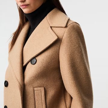 Nahaufnahme einer Frau im beigen Mantel mit breitem Revers und schwarzen Knöpfen – klassischer Look kombiniert mit schwarzem Rollkragenpullover.