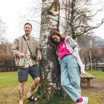 A man and a woman stand next to a tree, both leaning against it and smiling. The man holds a bottle and has a camera around his neck, and in the background there is a bench, a sign, and leafless trees.
