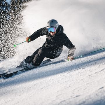 A skier in black ski clothing carves low across the slope, creating a large cloud of snow.