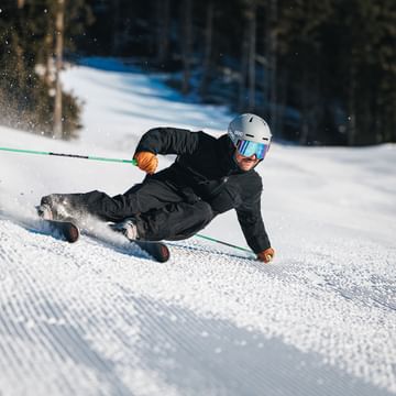 Ein Skifahrer fährt in tiefer Schräglage über eine präparierte Skipiste und wirbelt dabei Schnee auf. Im Hintergrund ist ein verschneiter Pistenabschnitt vor einem Wald zu sehen.