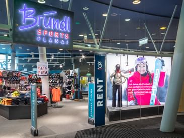 Das Bild zeigt den Eingang eines Bründl Sports Shops in der Planai mit einem großen, leuchtenden Bründl Sports Schild. Im Inneren sind verschiedene Wintersportprodukte wie Kleidung und Ausrüstung ausgestellt, und es gibt einen Bereich für Verleih und Service. Ein großes Werbeposter zeigt eine lächelnde Skifahrerin.