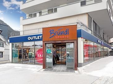 The image shows the entrance of a sports store with the sign “Bründl Sports” above the door. Large display windows show mannequins, and the words “OUTLET” and “SALE” are clearly visible.