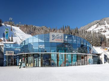 The image shows the Bründl Sports AreitXpress valley station shop with a large blue glass facade next to a snowy ski slope. The “Bründl Sports” logo and the texts “SKIDEPOT,” “SHOP,” “RENT,” “DEPOT,” and “SERVICE” are visible on the building, with a ski lift, forest, and snow-covered mountains in the background.