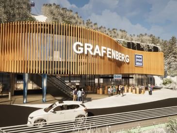 Ein modernes Gebäude mit geschwungener Holzfassade und dem Schriftzug „GRAFENBERG“ steht vor verschneiten Bäumen. Vor dem Eingang stehen mehrere Menschen, ein Auto fährt vorbei, und Schilder mit „Flachau Snow Space Salzburg“, „Bründl Sports“ sowie „Ticketschalter“ sind sichtbar, während oben eine Gondelbahn verläuft.