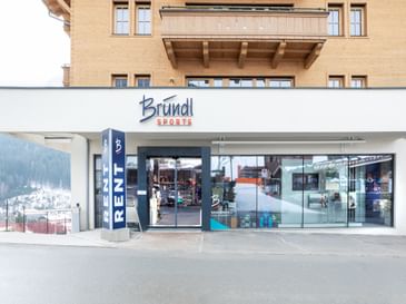 Das Bild zeigt den Eingang eines Bründl Sports Shops in einem alpinen Gebäude. Über dem Eingang befindet sich das Logo, und der Schriftzug "RENT" weist auf die Verleihmöglichkeit hin.